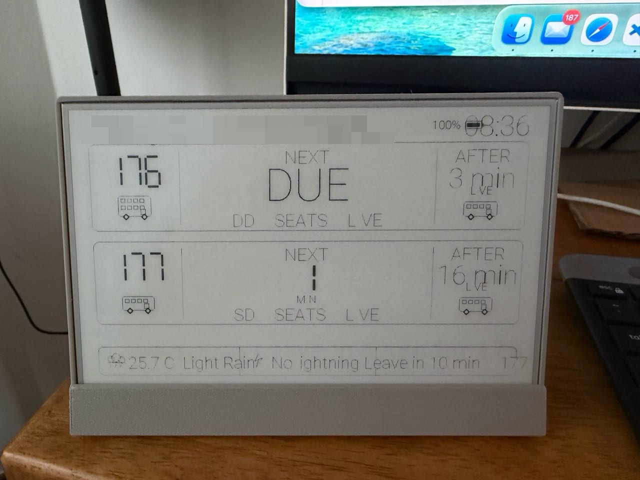 The screen in its stand showing a thin-lined layout with 176 due and 177 in one minute, a footer with the temperature and 'Light Rain', and visibly smudged, broken letters